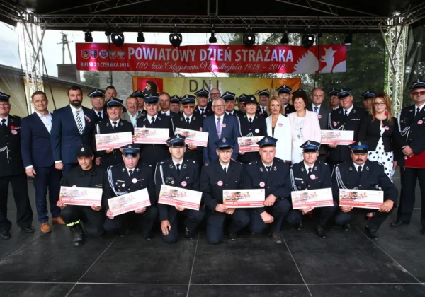 Grupa strażaków w mundurach stojących na scenie podczas Powiatowego Dnia Strażaka, prezentujących duże czeki, z banerem upamiętniającym 100-lecie Odzyskania Niepodległości w tle.