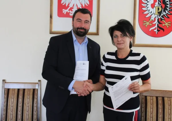 Mężczyzna w garniturze i kobieta w koszulce w paski ściskają dłonie po podpisaniu dokumentów, w tle widoczne herby powiatu płockiego, symbolizujące partnerstwo na rzecz wsparcia i rozwoju ekonomii społecznej.