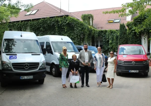 Grupa osób stoi przed nowymi pojazdami jednostek organizacyjnych powiatu płockiego: dwoma białymi vanami i czerwonym vanem marki Volkswagen, przed budynkiem porośniętym bluszczem.