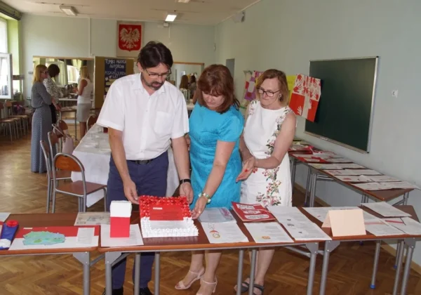 Trzy osoby, mężczyzna i dwie kobiety, kroją czerwony tort na stole pełnym dokumentów podczas zakończenia Powiatowego Konkursu Literackiego w Drobinie.