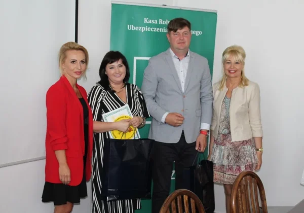 Cztery osoby stoją przed zielonym banerem Kasy Rolniczego Ubezpieczenia podczas rozstrzygnięcia konkursu „Bezpieczne Gospodarstwo Rolne 2018”, trzymając nagrody i torby.