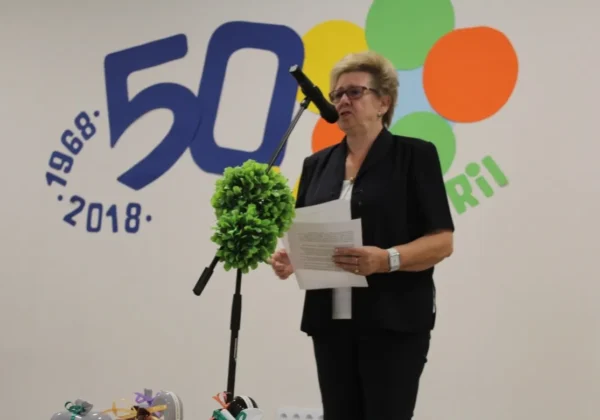 Kobieta w czarnym ubraniu stoi na scenie przy mikrofonie, trzyma kartki i przemawia podczas obchodów Światowego Dnia Inwalidy.