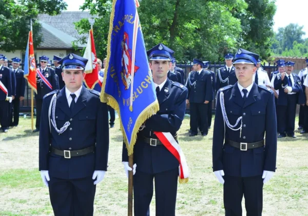 Podczas uroczystości 100-lecia Ochotniczej Straży Pożarnej w Niesłuchowie trzech młodych strażaków w mundurach z białymi rękawiczkami stoi na trawie, trzymając niebieski sztandar.