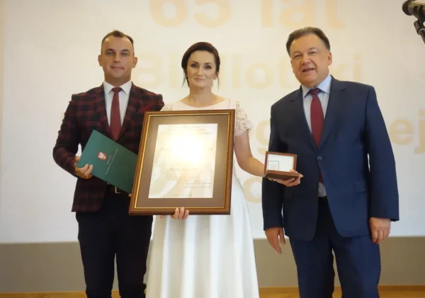 Trzy osoby stoją na scenie podczas 65-lecia Biblioteki Pedagogicznej w Płocku: mężczyzna w kratkowanej marynarce trzyma zielony folder, kobieta w białej sukni prezentuje duży oprawiony dyplom, a drugi mężczyzna w garniturze trzyma medal w pudełku.