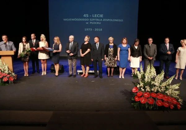 Grupa osób ubranych w formalne stroje stoi na scenie podczas jubileuszu 45-lecia Wojewódzkiego Szpitala Zespolonego w Płocku, z podiumem po lewej i dekoracjami z kwiatów na pierwszym planie.