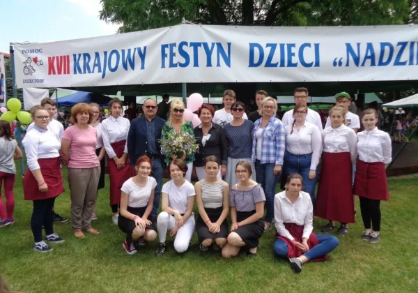 Grupa ludzi w różnym wieku stoi na trawie pod bannerem „KRAJOWY FESTYN DZIECI NADZIEJA”, podczas plenerowego festynu.