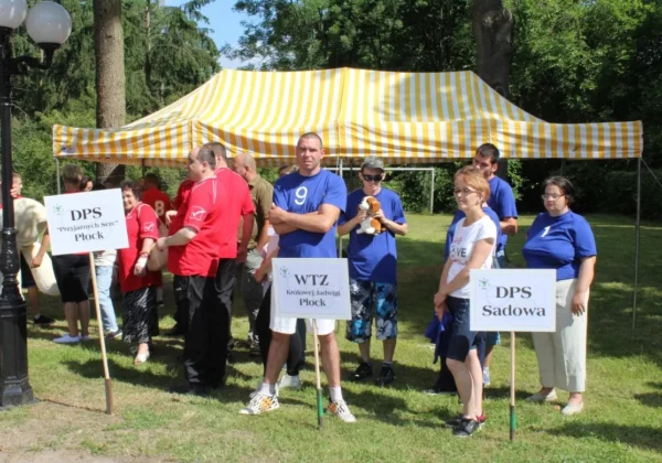 Uczestnicy XXVI Letniej Olimpiady Sportowej Osób Niepełnosprawnych w Nowym Miszewie stoją pod żółto‑białym namiotem, trzymając tablice z nazwami ośrodków.