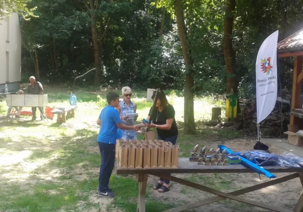 Na zewnątrz, przy drewnianym stole, młody uczestnik w niebieskiej koszulce odbiera nagrodę od dorosłej kobiety podczas Zawodów Wędkarskich Służb Mundurowych w Małej Wsi; na stole widać pakunki nagród i trofea w kształcie ryb, a w tle stoją banery i drzewa.