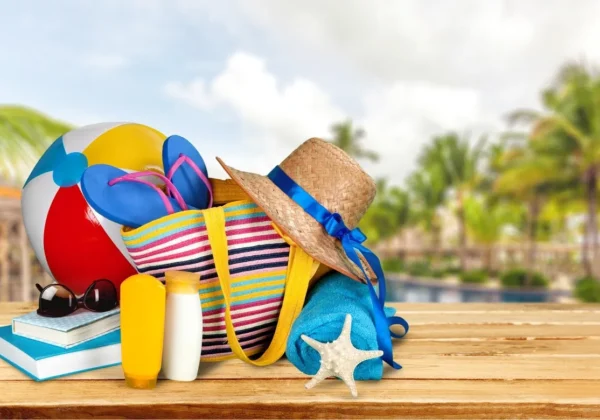 Kolorowa plażowa wyprawka na drewnianym blacie: torba z kapeluszem, okulary przeciwsłoneczne, kremy, książki, zabawki plażowe i gwiazdka morska, z palmami w tle.