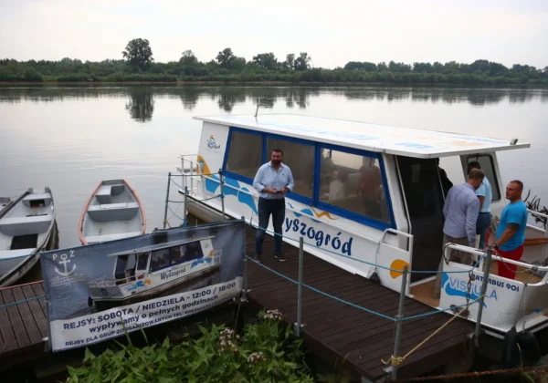 Wycieczkowy statek cumuje przy drewnianym molo nad rzeką Wisłą w Wyszogrodzie, a grupa osób spaceruje i rozmawia na pokładzie.