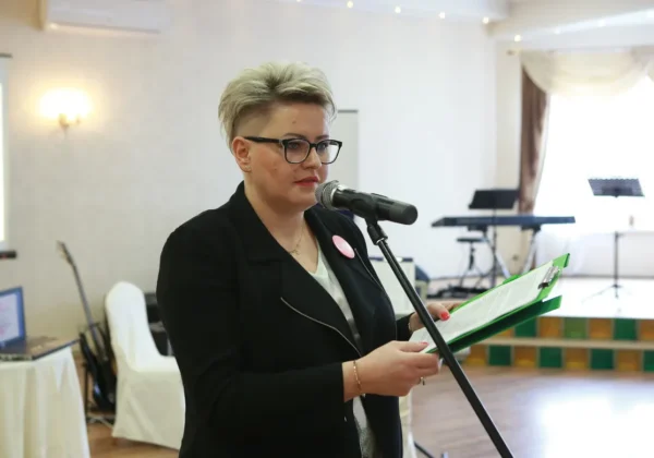 Kobieta w czarnym ubraniu stoi przy mikrofonie i trzyma zielony folder z dokumentami podczas wydarzenia Konkurs na Najaktywniejszą Liderkę Wiejską na Mazowszu.