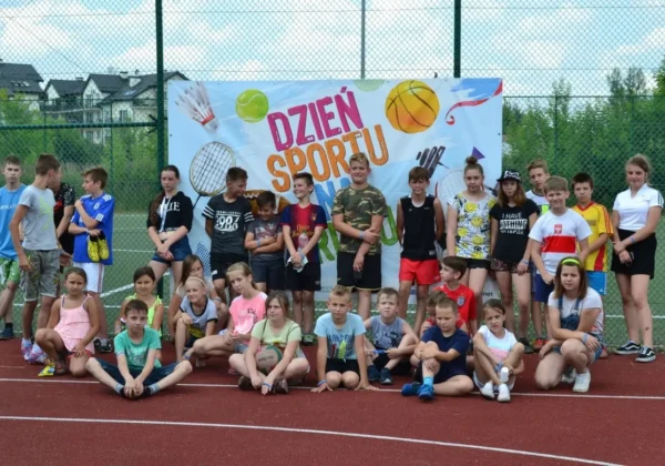 Grupa dzieci i młodzieży stoi na Orliku przed kolorowym banerem z napisem Dzień Sportu.