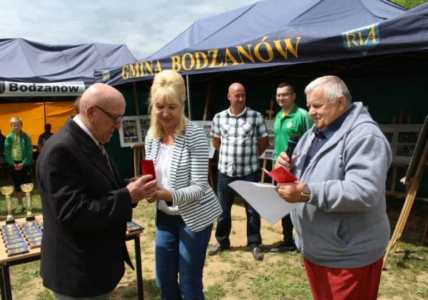 Podczas Historycznego Przeglądu Klubu Sportowego Huragan Bodzanów pod namiotem gminy Bodzanów, starszy mężczyzna w garniturze odbiera od blond kobiety nagrodę w czerwonym pudełku, podczas gdy inny mężczyzna z mikrofonem wygłasza przemówienie.