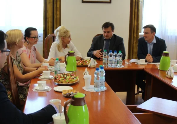 Grupa osób w formalnych ubraniach siedzi przy stole konferencyjnym i omawia porozumienie o współpracy w projekcie „Liderzy kooperacji”, z napojami i przekąskami na stole.