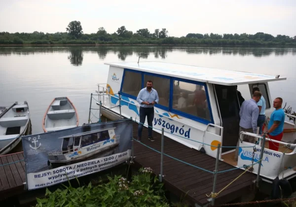 Wycieczkowa łódź motorowa z niebieskimi elementami stoi przy drewnianym pomoście na spokojnym jeziorze, a obok widoczne są osoby i banner informacyjny o rejsach.