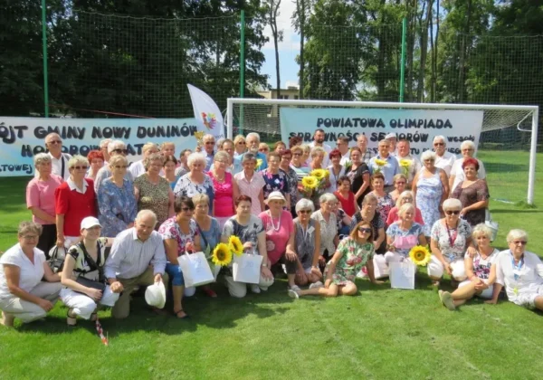 Grupa seniorów stojących i siedzących na zielonej trawie przed bannerami z napisem „Powiatowa Olimpiada Sprawności Seniorów na Wesoło” podczas wydarzenia o Puchar Starosty Płockiego.