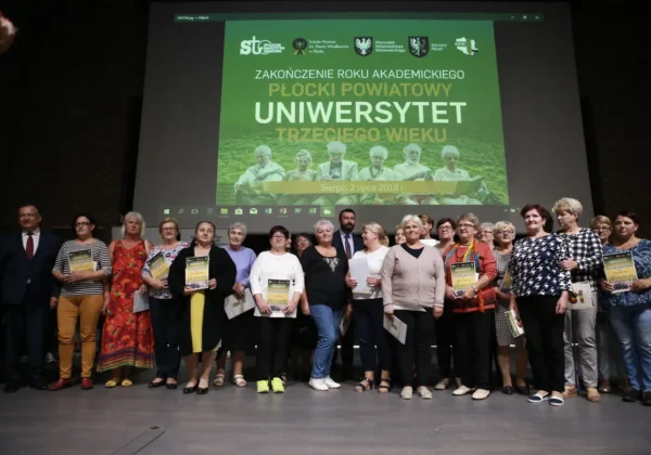 Grupa seniorów stoi na scenie podczas zakończenia roku akademickiego Powiatowego Uniwersytetu Trzeciego Wieku, trzymając certyfikaty i dyplomy przed wyświetlanym plakatem wydarzenia.