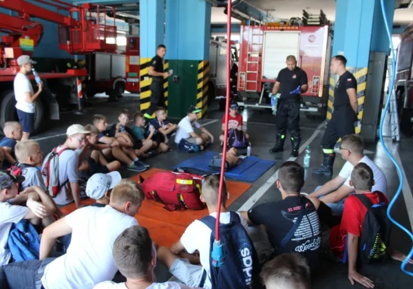 Grupa młodzieżowych drużyn pożarniczych siedzi na matach na podłodze remizy strażackiej, słuchając instruktorów podczas zajęć na obozie w 2018 roku.