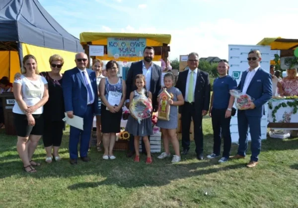 Grupa dorosłych i dzieci stoi na zielonej trawie podczas Dni Gminy Bodzanów, przed kolorowymi namiotami i stoiskami.