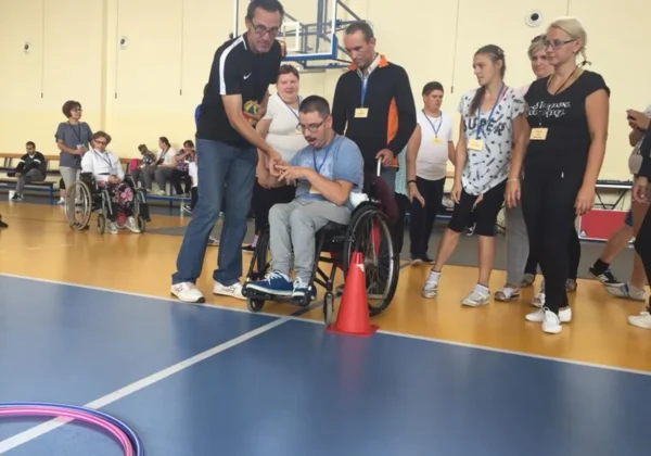 Podczas II Powiatowej Spartakiady Integracyjnej Osób Niepełnosprawnych w Radzanowie 2018, mężczyzna na wózku inwalidzkim odbiera medal od stojącego obok mężczyzny, w otoczeniu innych uczestników z identyfikatorami.