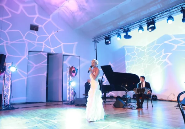 Kobieta w białej sukni śpiewa na scenie podczas koncertu „Mazowsze w sercu Niepodległej”, towarzyszą jej pianista przy fortepianie i akordeonista, a scenę oświetlają kolorowe projekcje na ścianach i suficie.