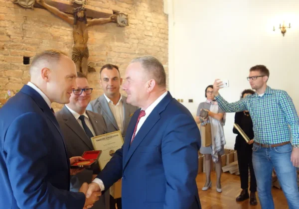 Dwóch mężczyzn w garniturach ściska sobie dłonie podczas ceremonii wręczania odznaczeń, w tle stoją inni uczestnicy, a na ceglanej ścianie wisi krzyż.