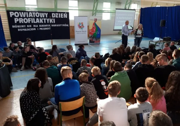 Duża grupa uczniów siedzi w sali gimnastycznej podczas Powiatowego Dnia Profilaktyki, na scenie trzech prowadzących przemawia, a w tle widoczny jest baner z hasłem „Mówimy NIE dla alkoholu, narkotyków, dopalaczy i przemocy”.