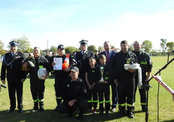 Grupa strażaków w mundurach bojowych wraz z innymi uczestnikami na boisku podczas Gminnych Zawodów Sportowo-Pożarniczych w Łącku, prezentujących trofeum i certyfikat.