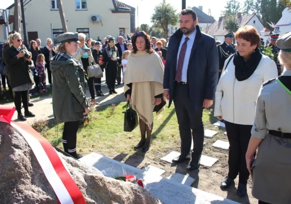 Grupa mieszkańców i urzędników uczestniczy w ceremonii przy kopcu z czerwono-białą wstęgą, podczas nadawania Parkowi Miejskiemu w Wyszogrodzie nowej nazwy.