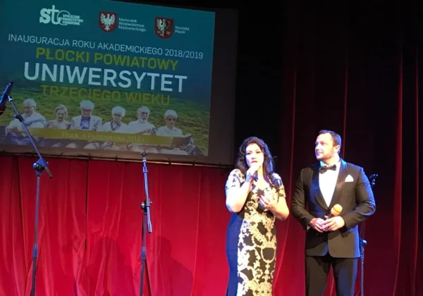 Dwie osoby w wieczorowych kreacjach stojące na scenie i mówiące do mikrofonów podczas II inauguracji Płockiego Powiatowego Uniwersytetu Trzeciego Wieku, z plakatem wydarzenia w tle.