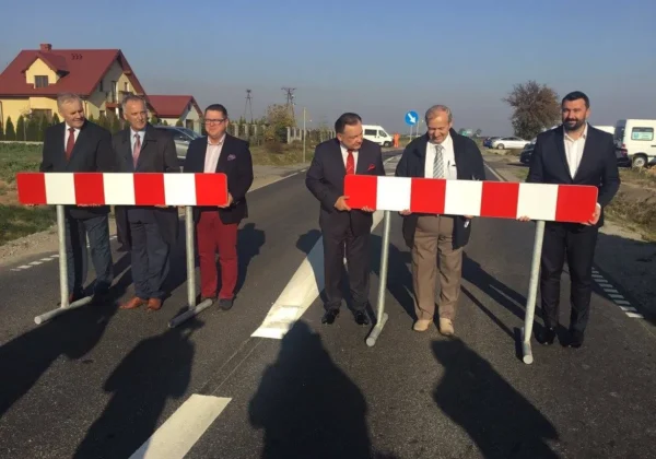 Grupa sześciu mężczyzn w garniturach stoi na nowo wybudowanym odcinku drogi w Gminie Bielsk, trzymając czerwono-białe bariery podczas otwarcia.