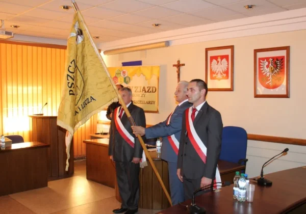 Trzech mężczyzn w garniturach ze biało‑czerwonymi wstążkami na piersiach stoi w sali sesyjnej płockiego Starostwa, a jeden z nich trzyma duży sztandar pszczelarski, w tle widoczne są godła i baner związany z pszczelarstwem.