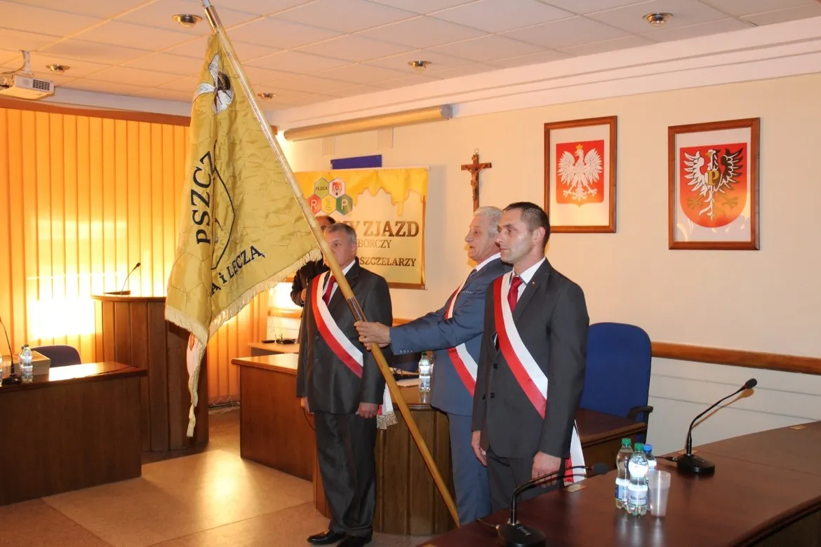 Trzech mężczyzn w garniturach ze biało‑czerwonymi wstążkami na piersiach stoi w sali sesyjnej płockiego Starostwa, a jeden z nich trzyma duży sztandar pszczelarski, w tle widoczne są godła i baner związany z pszczelarstwem.