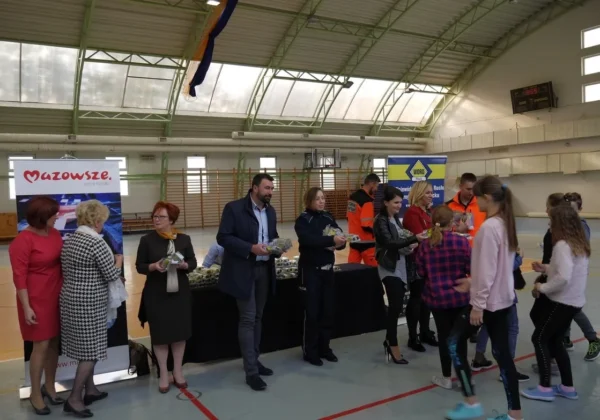 Uczniowie odbierają gadżety od dorosłych podczas programu „Jesteś widoczny, jesteś bezpieczny” w hali sportowej wyszogrodzkiej szkoły podstawowej, przy stoisku z banerami Mazowsze i WORD.