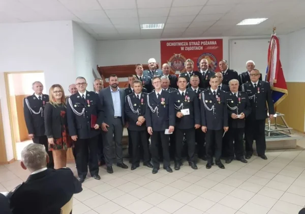Grupa druhów Ochotniczej Straży Pożarnej z Zągotach, Parzeniu i Bronowie Zalesiu w mundurach z odznaczeniami stojąca razem na sali podczas podsumowania działalności, z flagą w tle.