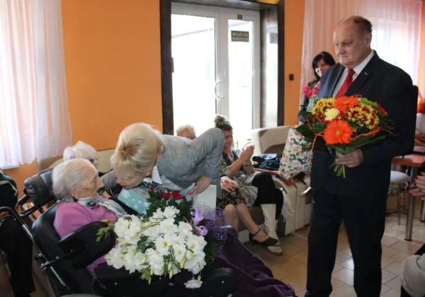 Mieszkanka domu pomocy społecznej w Koszelewie kończy 100 lat i otrzymuje bukiet kwiatów od mężczyzny w garniturze, podczas gdy inni seniorzy i goście jej gratulują.
