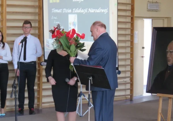 Podczas Dnia Edukacji Narodowej w Zespole Szkół w Gąbinie mężczyzna w garniturze wręcza kwiaty kobiecie na scenie, obok stoją uczniowie z mikrofonami, a po prawej stoi portret starszego mężczyzny na sztaludze.