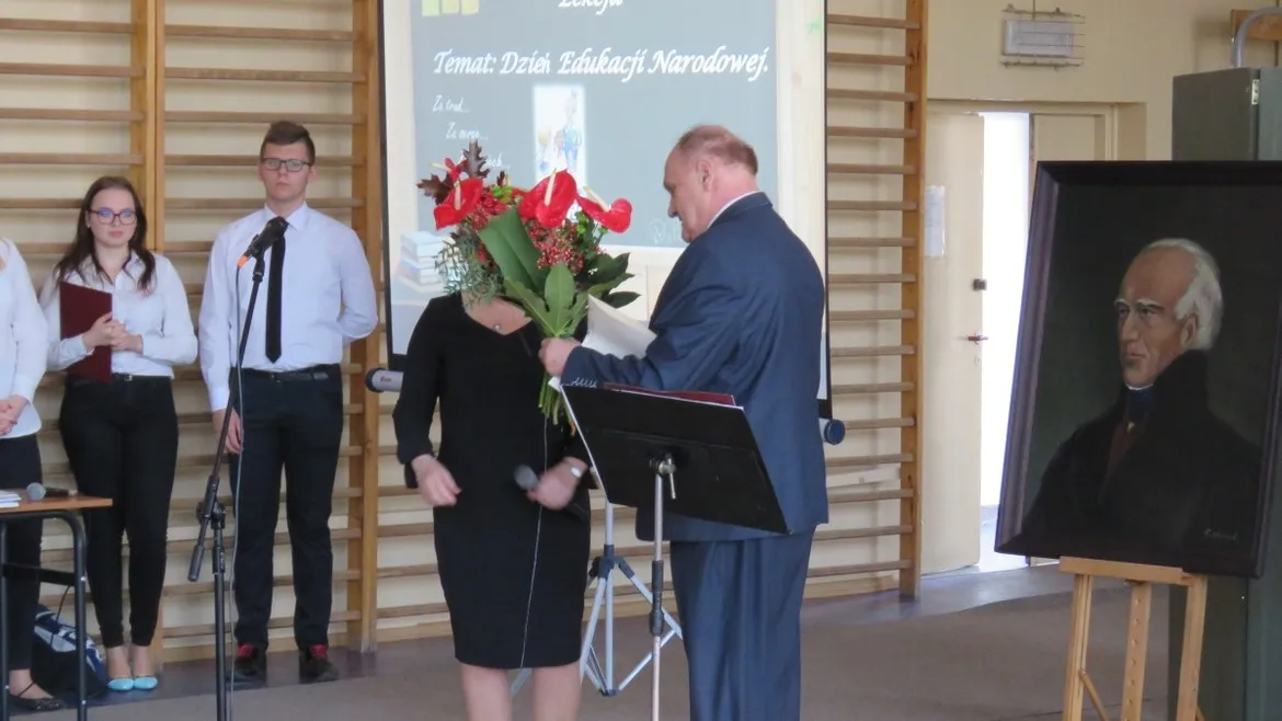 Podczas Dnia Edukacji Narodowej w Zespole Szkół w Gąbinie mężczyzna w garniturze wręcza kwiaty kobiecie na scenie, obok stoją uczniowie z mikrofonami, a po prawej stoi portret starszego mężczyzny na sztaludze.