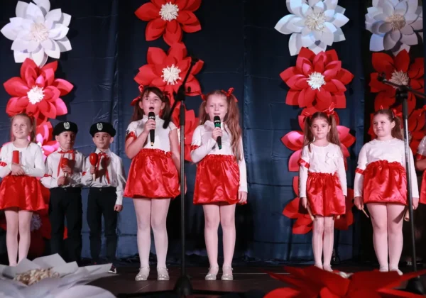 Dzieci w białych bluzkach i czerwonych spódniczkach stoją na scenie i śpiewają do mikrofonów podczas Dnia Edukacji Narodowej, z dekoracjami w tle w kształcie czerwono-białych kwiatów.