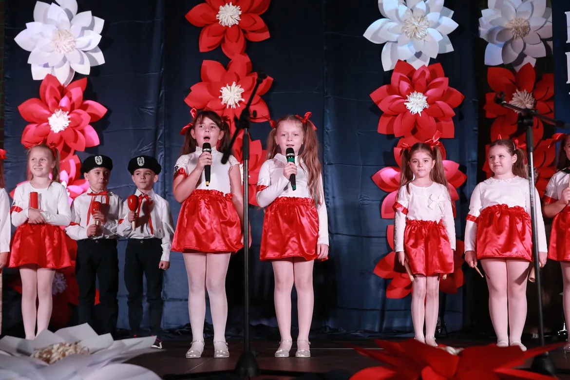 Dzieci w białych bluzkach i czerwonych spódniczkach stoją na scenie i śpiewają do mikrofonów podczas Dnia Edukacji Narodowej, z dekoracjami w tle w kształcie czerwono-białych kwiatów.