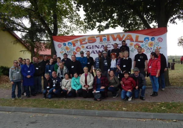 Grupa uczestników Festiwalu Ginących Zawodów w Wyszogrodzie stoi przed barwnym banerem z motywami kwiatowymi.