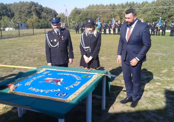 Uroczystość nadania sztandaru OSP w Szczytnie: trzy osoby w mundurach strażackich i garniturze stoją przy stole z błękitnym sztandarem ozdobionym frędzlami, w tle widoczna grupa druhów z chorągwiami.