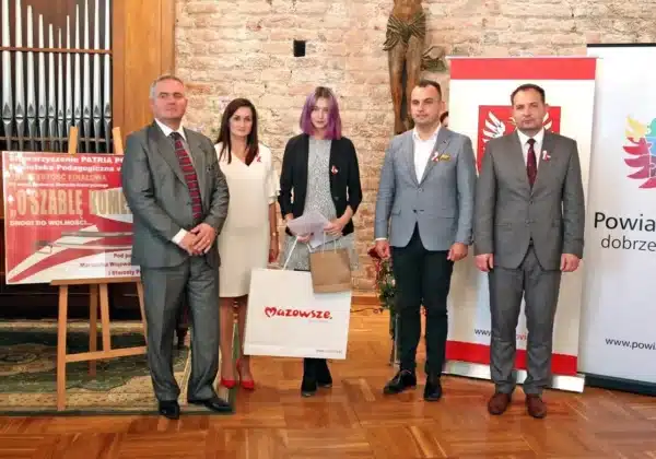 Na zdjęciu przedstawione jest pięć osób stojących w sali, z uśmiechami na twarzach, podczas wręczenia nagród w konkursie „O szablę Komendanta”, obok widoczne plakaty związane z wydarzeniem.