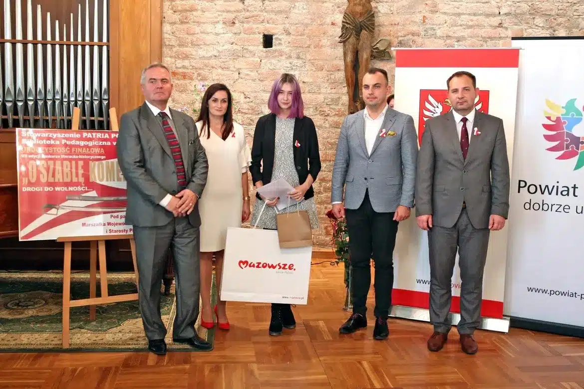 Na zdjęciu przedstawione jest pięć osób stojących w sali, z uśmiechami na twarzach, podczas wręczenia nagród w konkursie „O szablę Komendanta”, obok widoczne plakaty związane z wydarzeniem.