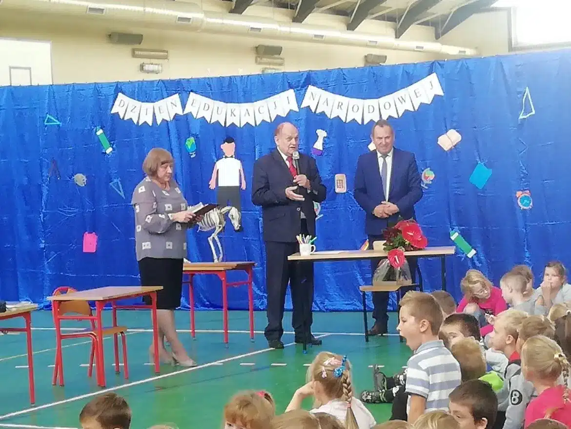 Uroczystość z okazji Dnia Edukacji Narodowej w Gminie Łąck, w której uczestniczą nauczyciele oraz dzieci zgromadzone na sali.