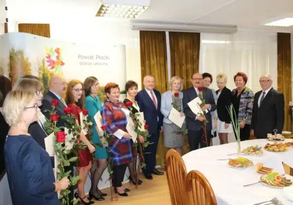 Grupa nauczycieli i przedstawicieli lokalnych władz świętuje XX Powiatowy Dzień Edukacji Narodowej, trzymając róże i dyplomy w eleganckim wnętrzu.