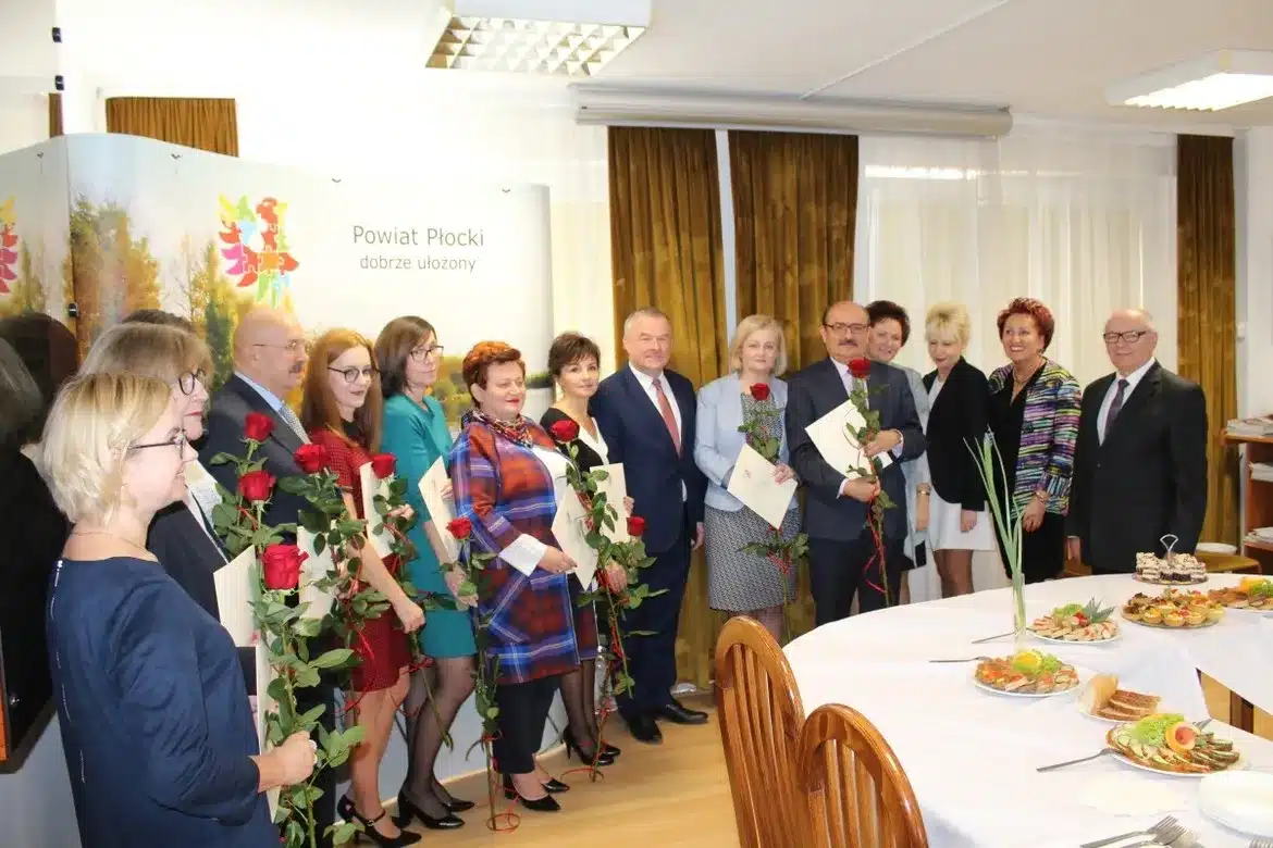 Grupa nauczycieli i przedstawicieli lokalnych władz świętuje XX Powiatowy Dzień Edukacji Narodowej, trzymając róże i dyplomy w eleganckim wnętrzu.