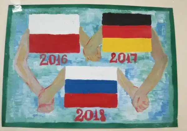 Na obrazie przedstawione są trzy pary rąk trzymające flagi Polski, Niemiec i Rosji, symbolizujące współpracę międzynarodową w latach 2016, 2017 i 2018.