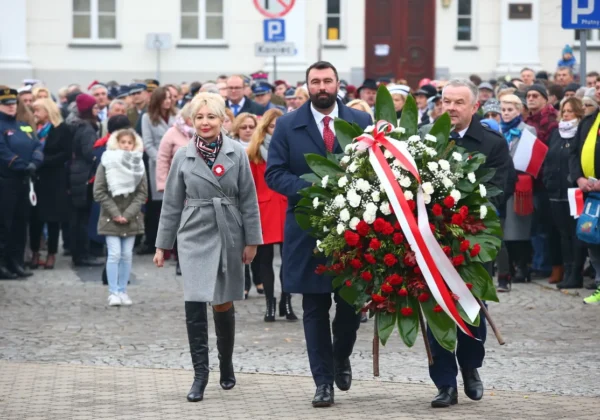 Para prowadząca uroczystość niesie duży wieniec z czerwonych i białych kwiatów ozdobiony wstęgami podczas obchodów 100-lecia niepodległości, w otoczeniu zgromadzonych uczestników.