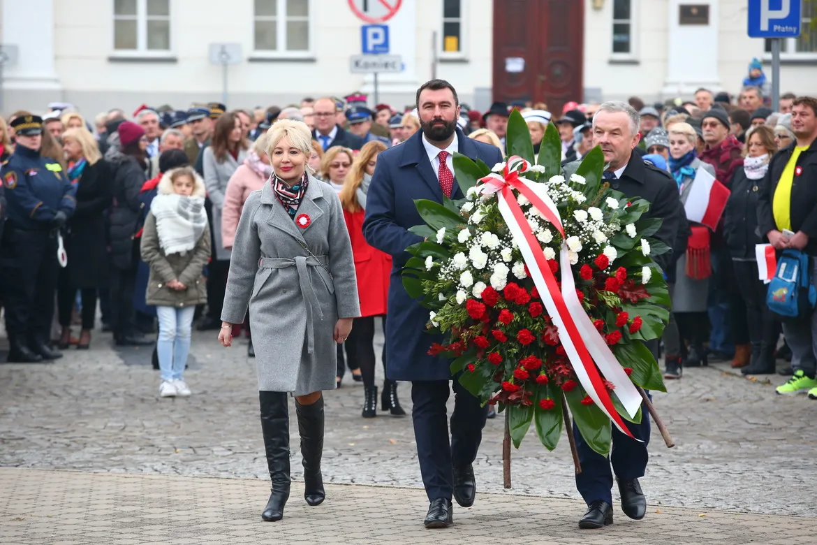 Para prowadząca uroczystość niesie duży wieniec z czerwonych i białych kwiatów ozdobiony wstęgami podczas obchodów 100-lecia niepodległości, w otoczeniu zgromadzonych uczestników.